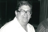 Un homme corpulent avec des lunettes épaisses et carrées sourit.