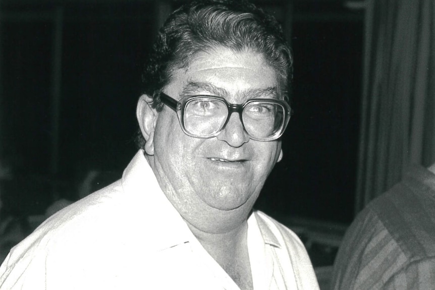 A portly man with thick, squarish glasses smiles.