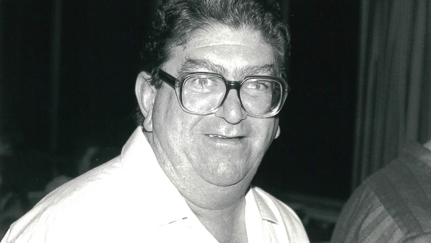 A portly man with thick, squarish glasses smiles.