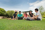Five young adults sitting on grass talking at a university campus.