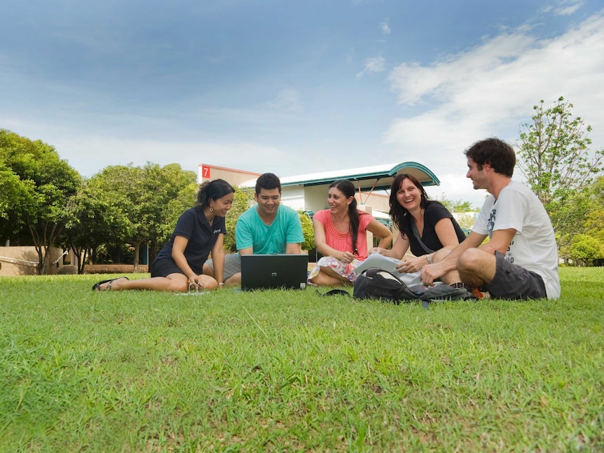 Five young adults sitting on grass talking at a university campus.
