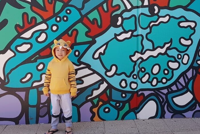 A little boy in a yellow hoodie stands smiling in front of a colourful mural of a crab.