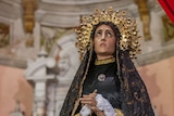 A statue of the Virgin Mary with olive skin looks up in dismay while adorned with a gold crown of stars and a black lace veil.