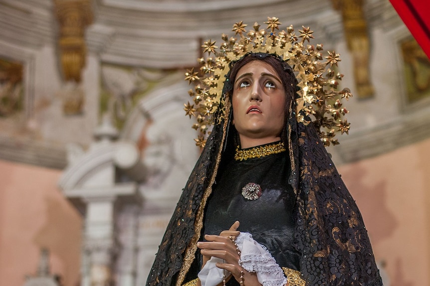 A statue of the Virgin Mary with olive skin looks up in dismay while adorned with a gold crown of stars and a black lace veil.