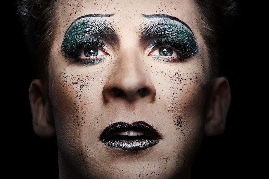 Close-up of a man's glittered face with heavy make-up and eyes looking upwards.