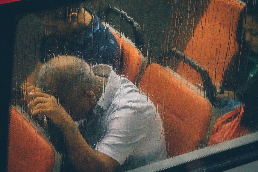 A rainy bus window and inside a man leaning his head on the seat in front of him.