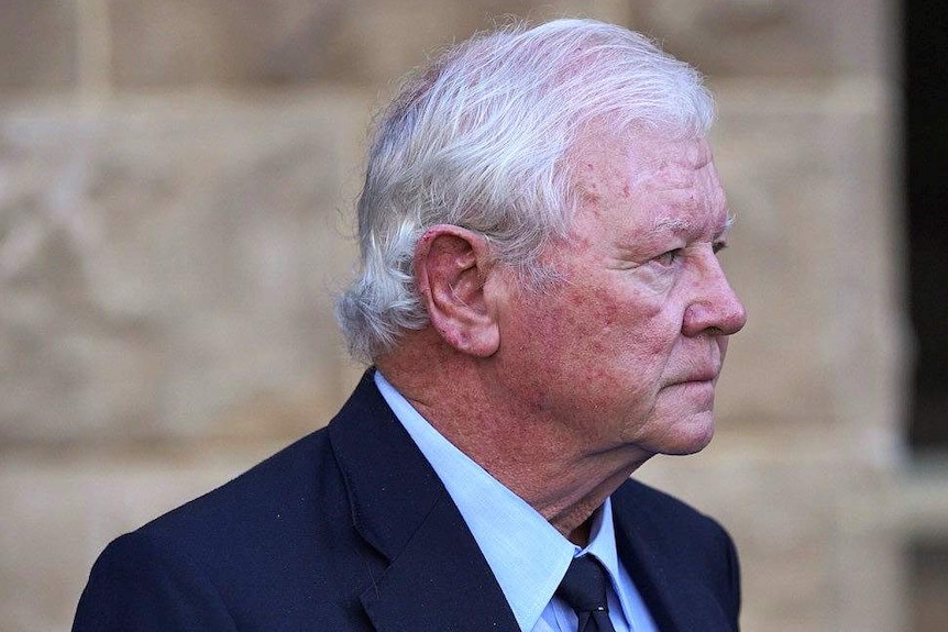 An older man wearing a dark blue suit at the front of a court building.