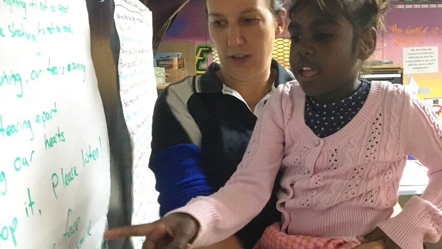 Alba Brockie holds an Indigenous girl in a pink cardigan, who points at a white board