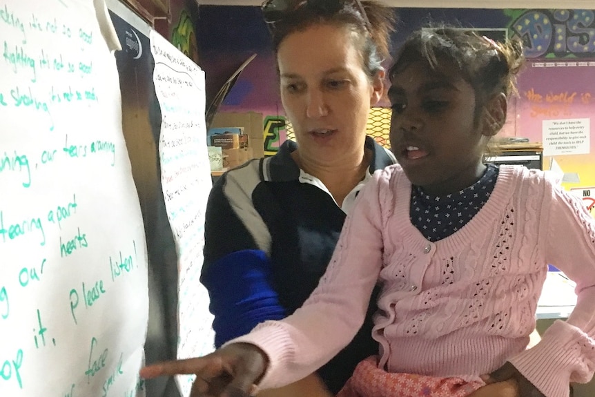 Alba Brockie holds an Indigenous girl in a pink cardigan, who points at a white board