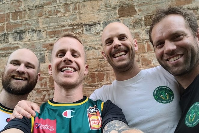 four smiling young men in basketball merchandise are in a selfie together in front of an old brick wall