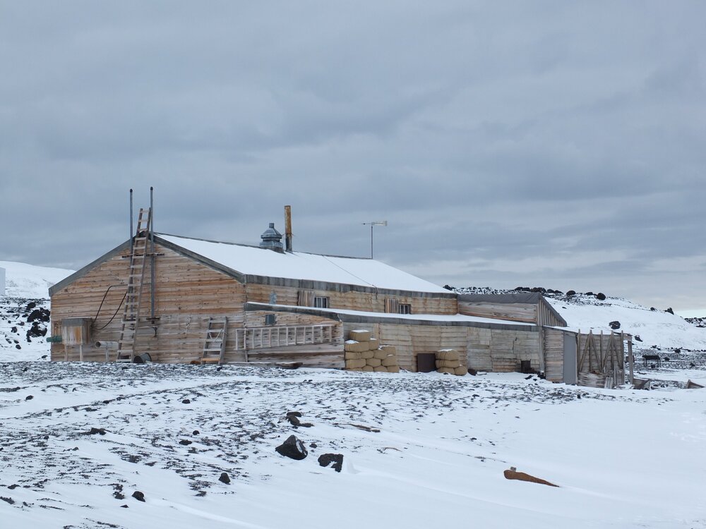 The long wooden structure, Scotts Terra Nova Hut, is situated in a snowy landscape, its roof dusted with snow.