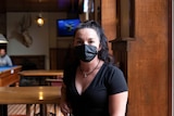 woman with dark hair wearing a mask sits at a table at a pub
