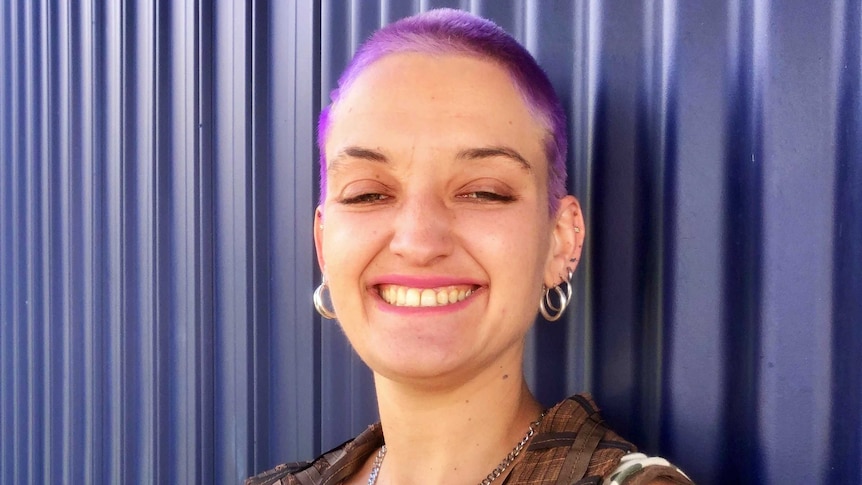A smiling young woman with short-cropped hair died bright purple, stands in front of a purple corrugated wall.