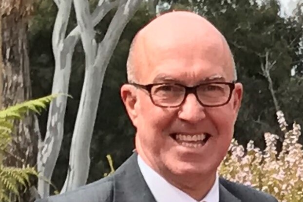 A man in a suit with a bald head smiles at the camera while wearing glasses.
