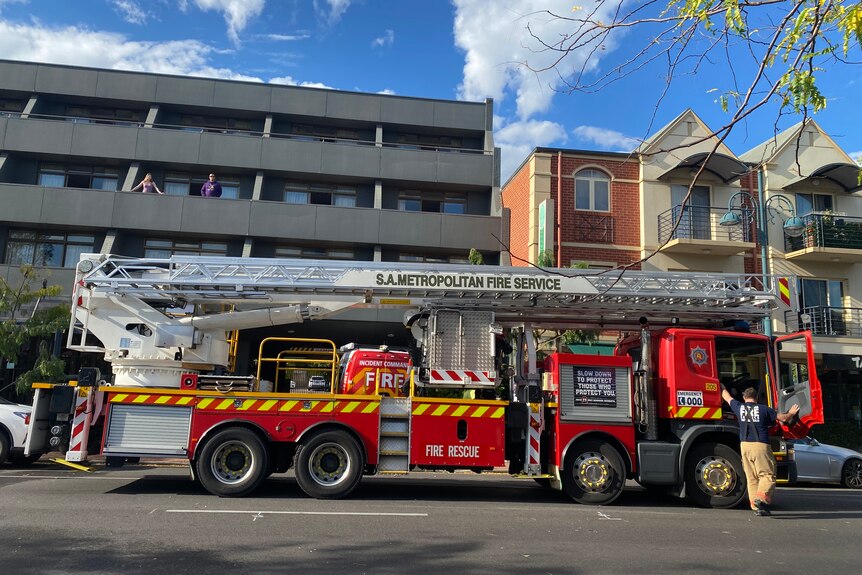 People stand on balconies in a modern building with a fire truck outside