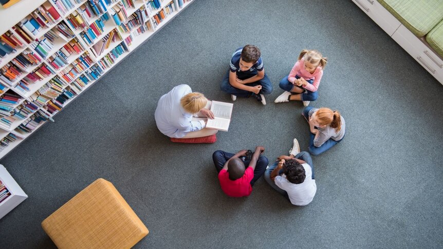 Top view of teacher reading book to children sitting in a circle on library floor.