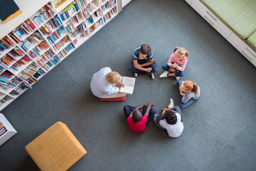 Top view of teacher reading book to children sitting in a circle on library floor.