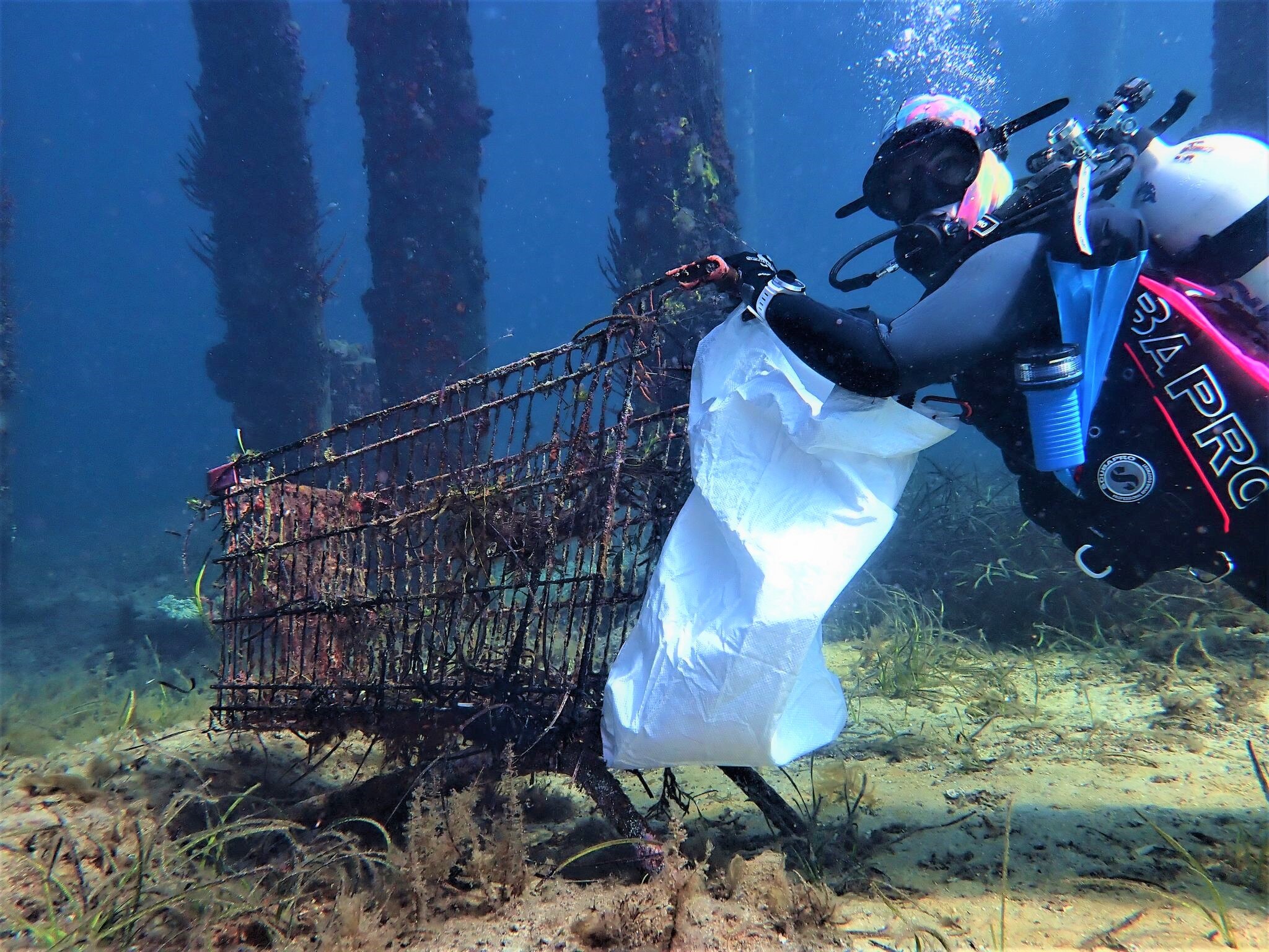 A scuba diva with a white bag attached to her wrist underwater holds on to the rails of a rusted trolley.