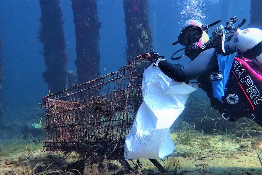 A scuba diva with a white bag attached to her wrist underwater holds on to the rails of a rusted trolley.