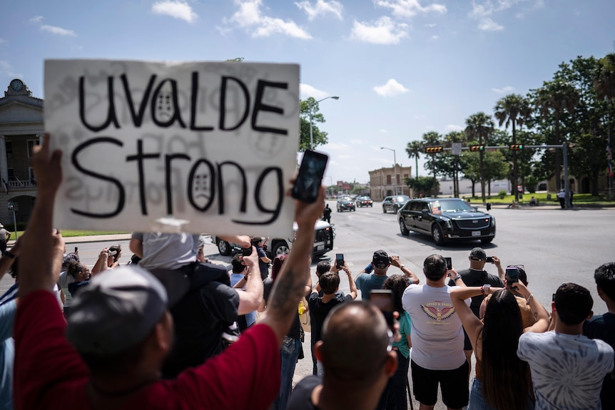 A black limo with a US flag on the front drives past a crowd of people, one man holds a sign that reads 'Uvalde Strong'