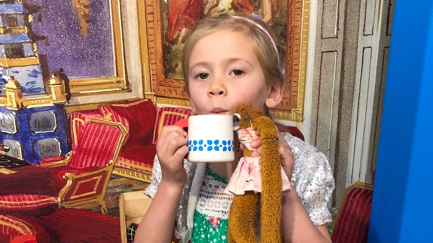 A girl holding a mug and a toy monkey in front of gilt-framed paintings