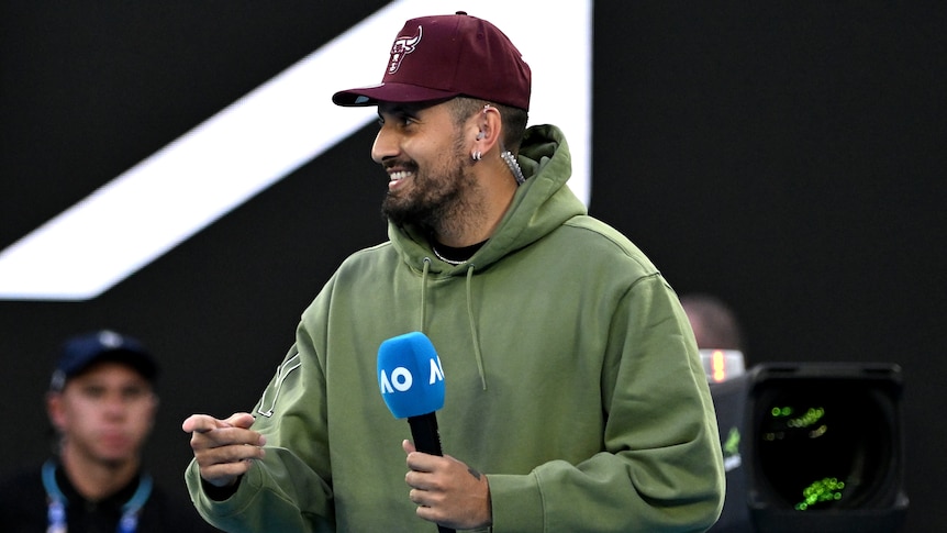 Nick Kyrgios smiles while holding a microphone on court before interviewing a player at the Australian Open.