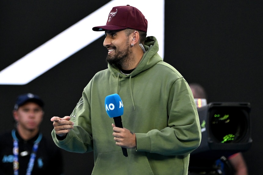 Nick Kyrgios smiles while holding a microphone on court before interviewing a player at the Australian Open.