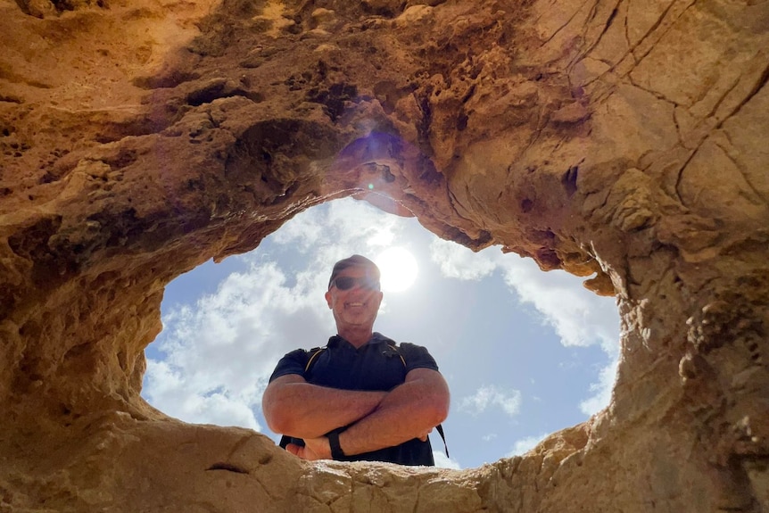 A man looking down into a hole he is smiling with crossed arms and a blue cloudy sky behind him.