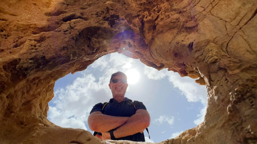 A man looking down into a hole he is smiling with crossed arms and a blue cloudy sky behind him.