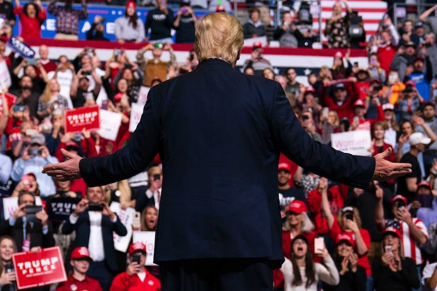 Donald Trump stands in front of fans, hands stretched out.