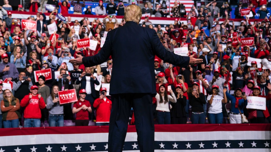 Donald Trump stands in front of fans, hands stretched out.