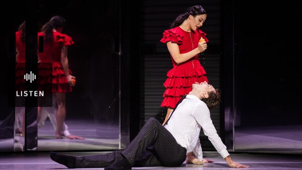 On stage, a woman in a red dress squeezes the juice of an orange into the mouth of a man sitting on the floor. Has Audio.