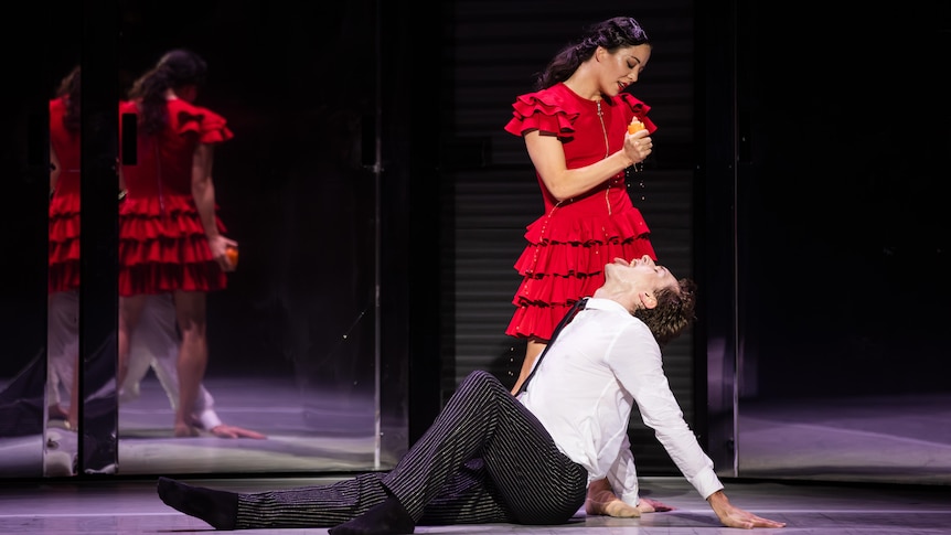 On stage, a woman in a red dress squeezes the juice of an orange into the mouth of a man sitting on the floor.