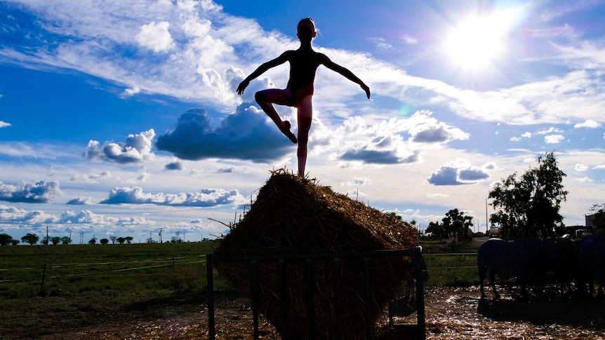 A young girl perches on a hay bale in a feeder, posing as a ballerina with the sun behind her.