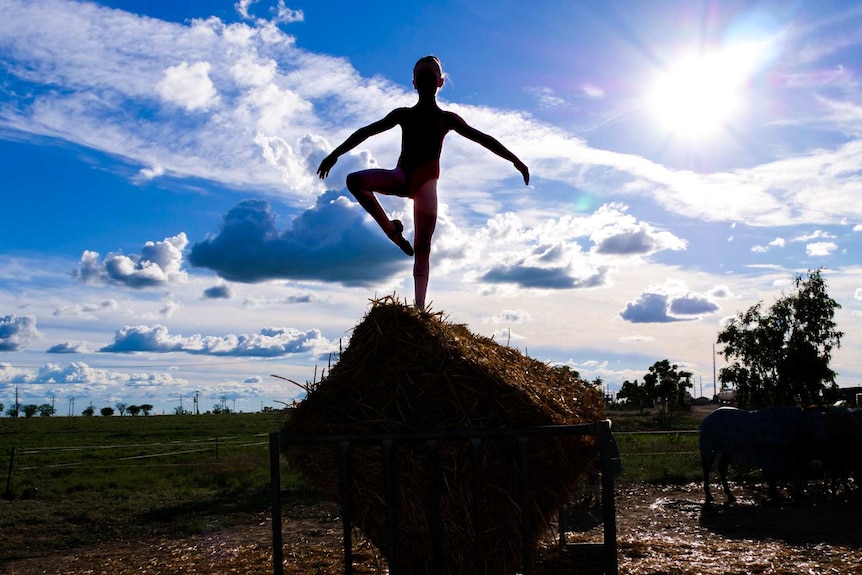 A young girl perches on a hay bale in a feeder, posing as a ballerina with the sun behind her.