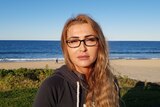 Aneta Duncan, wearing a dark hoodie top and black-rimmed glasses, sitting with a beach and ocean in the background.