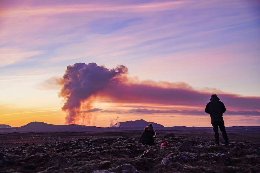 Two people seen in silhouette watch a rising plume of smoke from a volcanic eruption in the distance.
