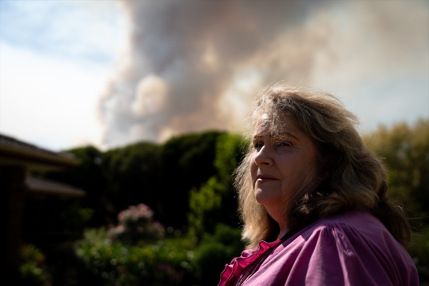 A woman stands in the foreground with a big smoke plume behind her