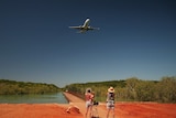 Two women and two children stand in red earth front of a pier, watching a plane soaring overhead.