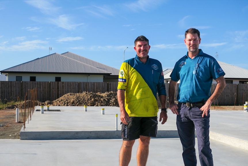 Builders, Cameron McInnes and Rob Rule stand opposite each other on a concrete building foundation with finished homes behind.