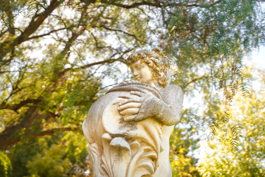 An old water fountain monument of a woman under a leafy pepper tree. 