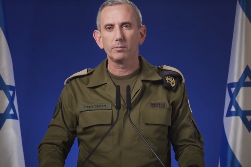 A man in a military uniform stands in from of two Israeli flags. 