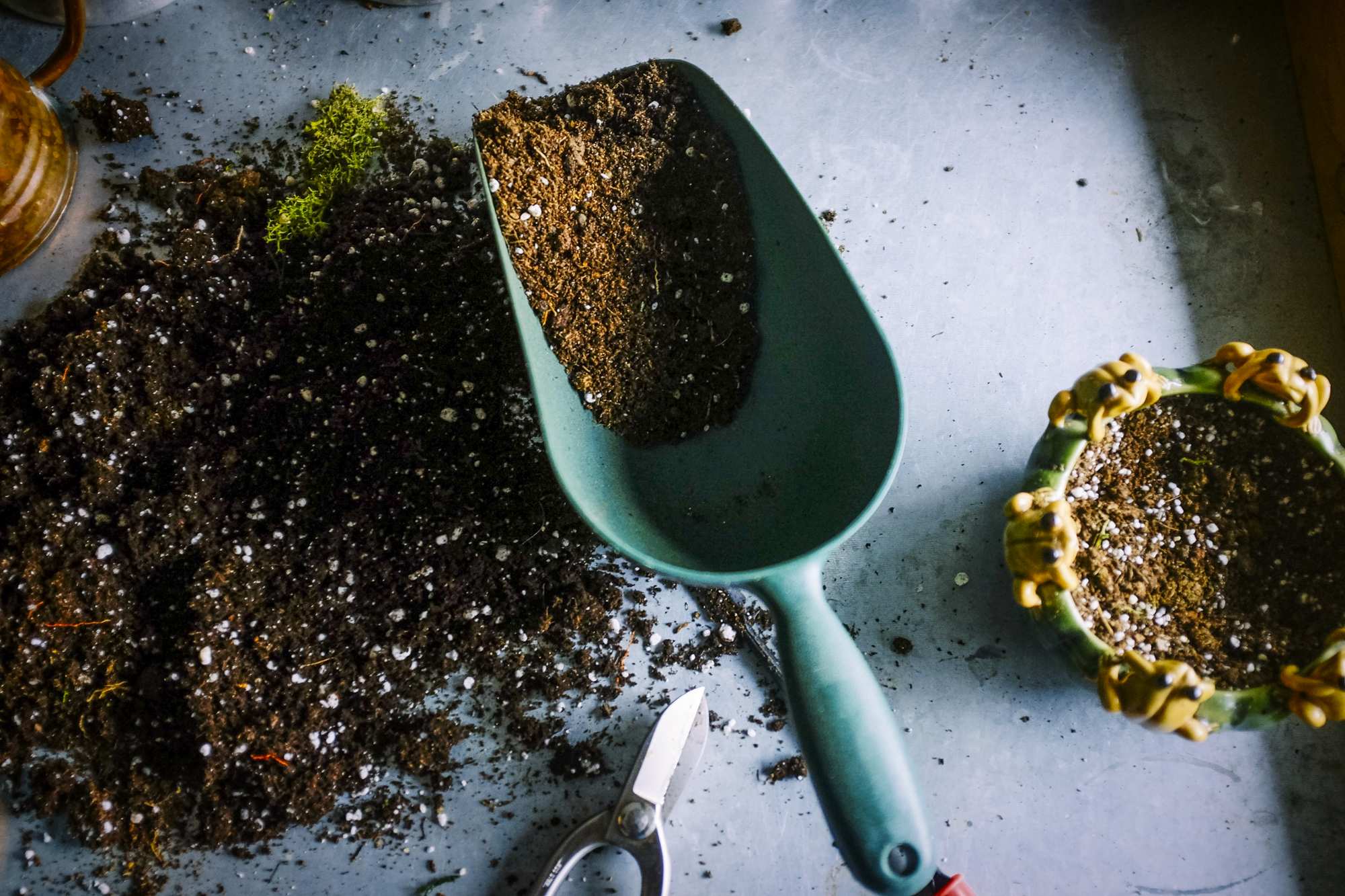 A trowel, half full of dirt, sitting on a metal bench, with dirt and plant pot beside it.