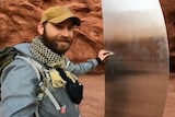 A man wearing hiking gear presses a small magnet onto the side of a tall shiny metal object among brown and orange desert.