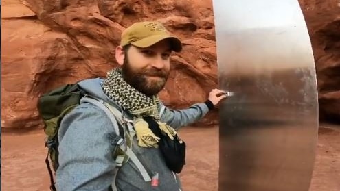 A man wearing hiking gear presses a small magnet onto the side of a tall shiny metal object among brown and orange desert.