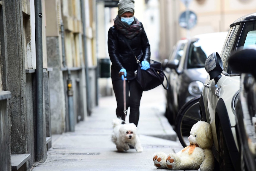 A woman wearing a face mask and winter clothes walks her dog on footpath on a deserted street.