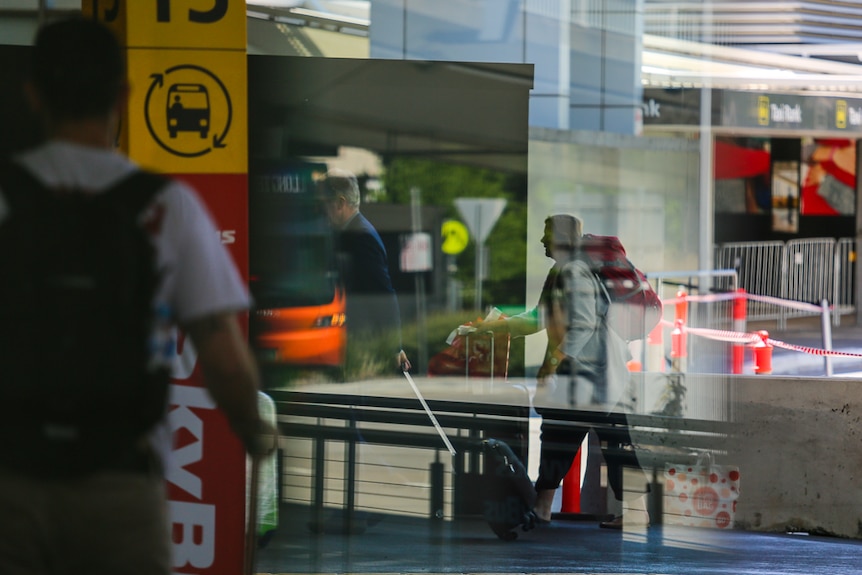 Travellers wearing packs and wheeling suitcases into Melbourne's Tullamarine Airport on a sunny day.