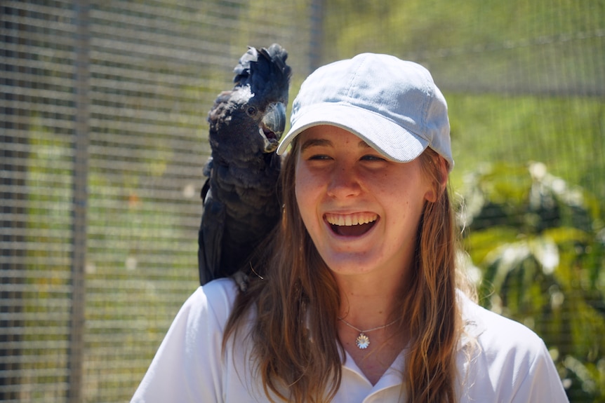 A young girl wearing a white polo shirt and white hat with a black cockatoo on her shoulder