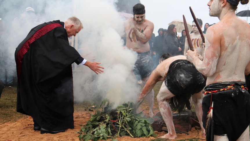 A judge in robes bends to take smoke from fire surrounded by Indigenous dancers