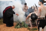 A judge in robes bends to take smoke from fire surrounded by Indigenous dancers